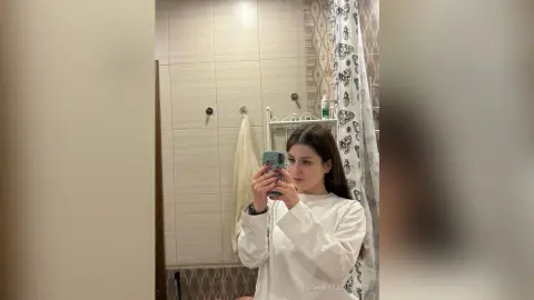 Young woman in a white long-sleeve shirt taking a mirror selfie in a modern tiled bathroom.