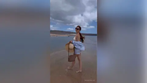 Stylish woman in a white swimsuit and blue cover-up walking on a serene beach under a cloudy sky.