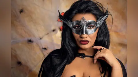 MissyJolie wears a silver horned mask and black lace outfit against a spiderweb background.