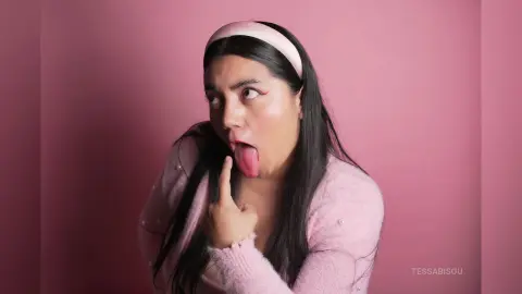 Playful young woman with long dark hair in a pink sweater and headband making a funny face against a pink background.