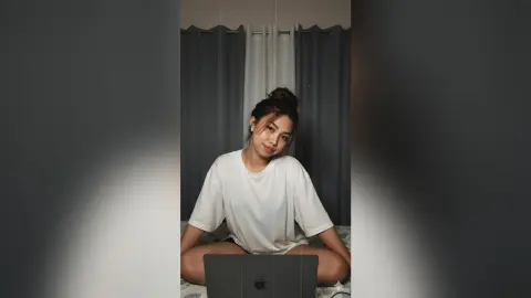 Young woman in a white t-shirt sitting with a laptop in a dimly lit room, creating a cozy study mood.