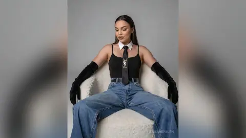 Elegant woman in black bodysuit, tie, and gloves sitting on a white chair in a minimalist studio setting.