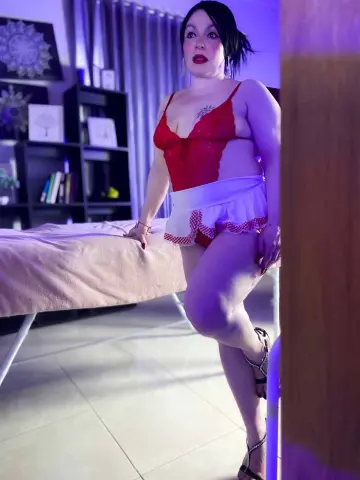 Woman in red lace bodysuit and white ruffled skirt sitting on a bed in moody purple lighting.