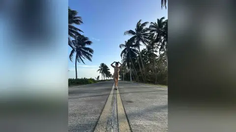 JulianaNavarro stands naked on a long road lined with palm trees, posing with her hands behind her head against a clear blue sky.