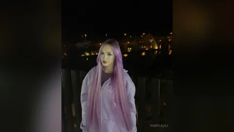 Young woman with long purple hair wearing a lavender hoodie at a night waterfront setting.