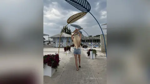 Blonde woman in denim shorts walking through a decorated beach resort path under a cloudy sky.
