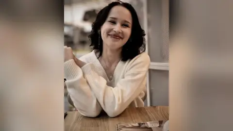 Smiling woman in a cozy white knit sweater sitting at a wooden table in a bright indoor setting.