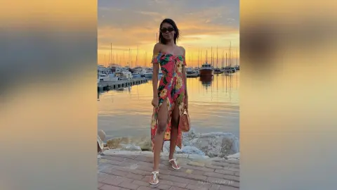 Stylish woman in a floral maxi dress posing by a harbor at sunset.