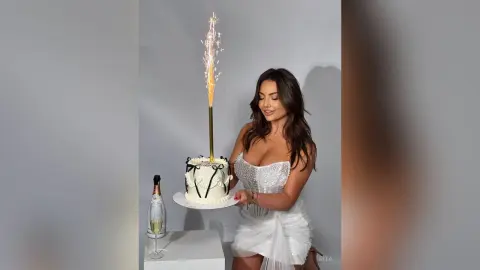 Smiling woman in a white sequin dress holding a birthday cake with a sparkling candle in a studio setting.