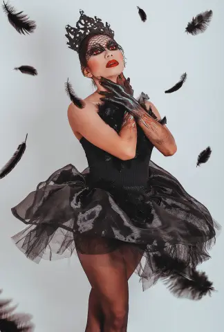 Elegant woman in black gothic tutu and crown surrounded by floating feathers in a moody studio setting.