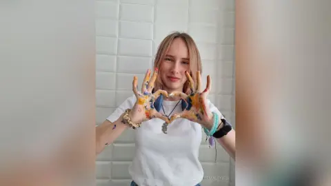 LisetteMaupredi smiles while holding her hands up in a heart shape, showing off her hands covered in colorful paint against a white tiled wall.