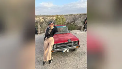 AlanaPearce poses next to a red car in a desert landscape, wearing a black long-sleeve top, tan pants, and a black baseball cap.