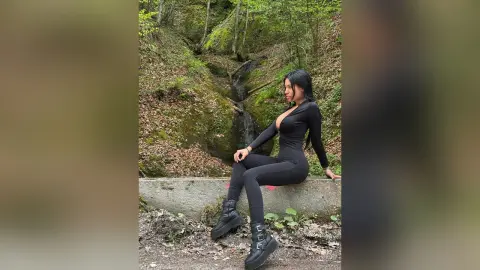 Woman in a black long-sleeve bodysuit sitting on a stone ledge in a lush green forest setting.