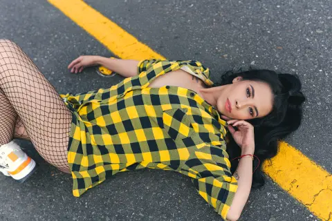 Young woman in a yellow plaid shirt and fishnet tights lying on an asphalt road with yellow lines.