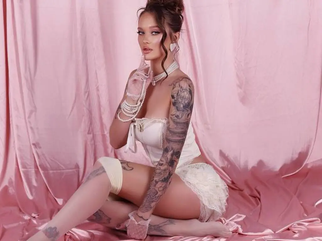 UliKuper poses in white lace lingerie against a pink satin backdrop, showing off her tattoos and pearl jewelry.