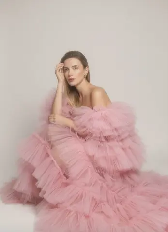 Elegant woman in a voluminous pink ruffled tulle gown posing against a neutral studio background.
