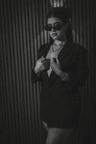 Elegant woman in a black blazer and sunglasses posing against a dark textured wall in a moody black and white portrait.