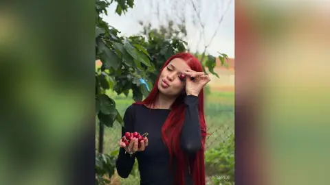 AriaVex holds a handful of red cherries in front of her black long-sleeved top, standing outdoors near green leaves.