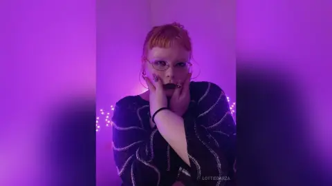 Redheaded woman in a black patterned sweater posing under moody purple neon lighting.