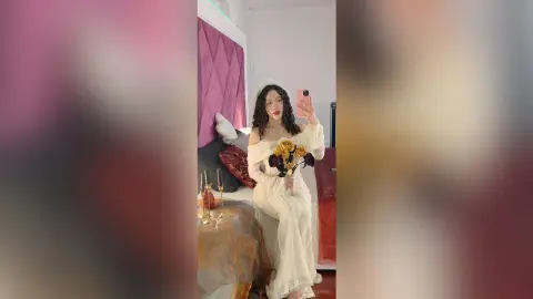 Elegant woman in a white lace wedding dress holding a bouquet in a romantic bedroom setting.