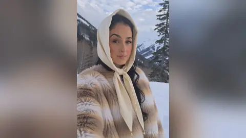 Smiling woman in a cream headscarf and faux fur coat enjoying a snowy mountain landscape.