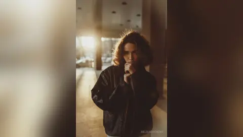 Moody portrait of a woman in a black leather jacket standing in a sunlit, modern hallway.