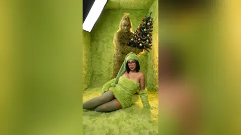 Woman in green fuzzy outfit sitting near Grinch character and Christmas tree in a festive lime green studio.