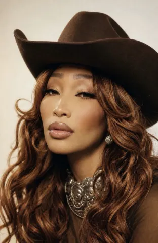 SOBLU — SOBLU wears a brown cowboy hat and a large silver necklace while looking into the camera with long wavy hair.