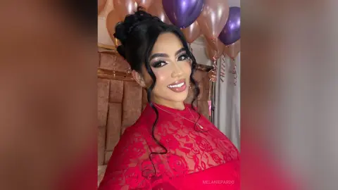 Smiling woman in a red lace dress celebrating at a party with purple and gold balloons.