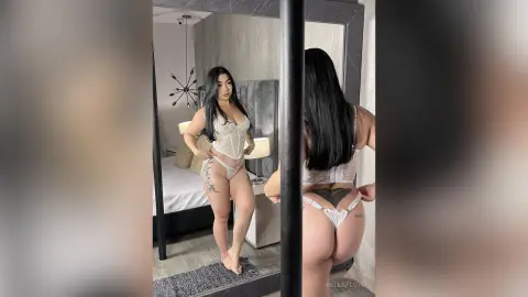 Young woman in white lace lingerie posing in a modern bedroom mirror for a glamorous lifestyle shot.