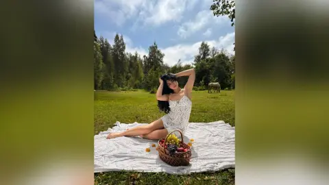 MinnieMile sits on a white blanket in a grassy field, wearing a white lace top and posing with one hand behind her head.