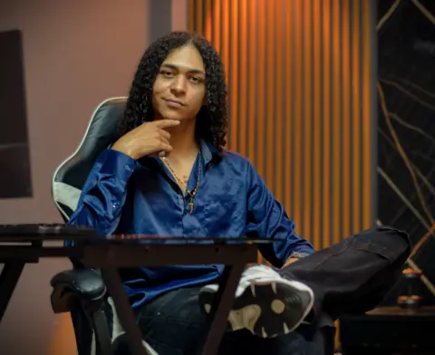 KylePresly sits in a black gaming chair wearing a blue satin button-down shirt and black pants.