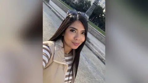 Smiling woman in a beige puffer vest and striped shirt posing outdoors on a sunny city sidewalk.