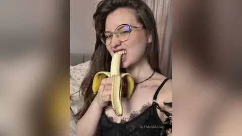 Young woman with glasses in a black lace camisole eating a banana in a bright indoor setting.