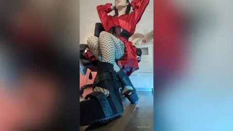 Woman in a red dress, fishnet tights, and chunky black platform sandals sitting in a modern room.