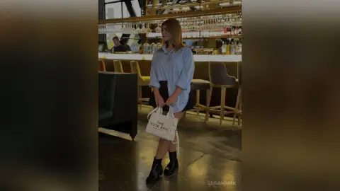 Young woman in a blue button-down shirt and black skirt holding a white tote bag in a modern dimly lit bar.