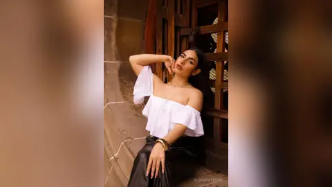 AlmaWhitmore poses with one hand behind her head, wearing an off-the-shoulder white top and a black skirt against a stone wall and wooden door.