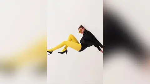 Stylish woman in yellow leggings and black top posing playfully against a white background.