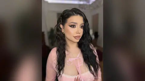 Young woman with long wavy hair and winged eyeliner wearing a pink lace top in a soft indoor setting.