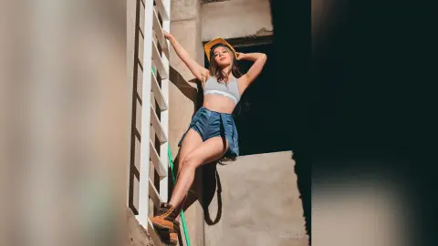 JessieLiu poses leaning against a white wall in a grey crop top and blue denim shorts.