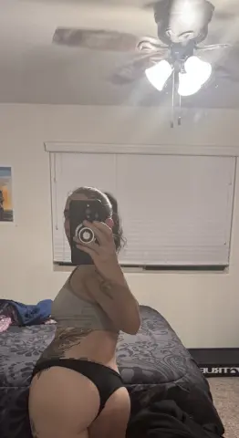 Woman in grey sports bra and black bikini briefs taking a mirror selfie in a dimly lit bedroom.