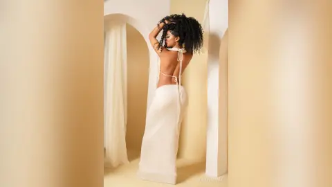 Elegant woman in a backless white maxi dress posing in a minimalist cream arched studio setting.