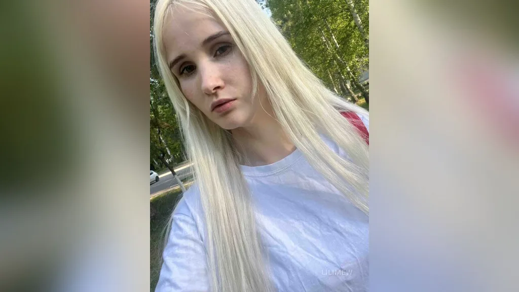 LiliMew — LiliMew with long blonde hair wears a white t-shirt while posing outdoors near green trees.