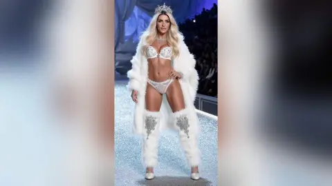 JessycaPage poses in white sparkly lingerie, a white fur coat, and a silver crown on a runway.
