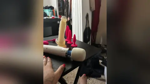 Assorted pink and flesh-toned adult sex toys arranged on a black table in a bedroom setting.