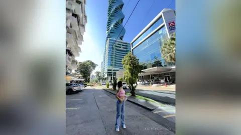Young woman in a pink crop top and jeans standing on a sunny city street near modern skyscrapers.