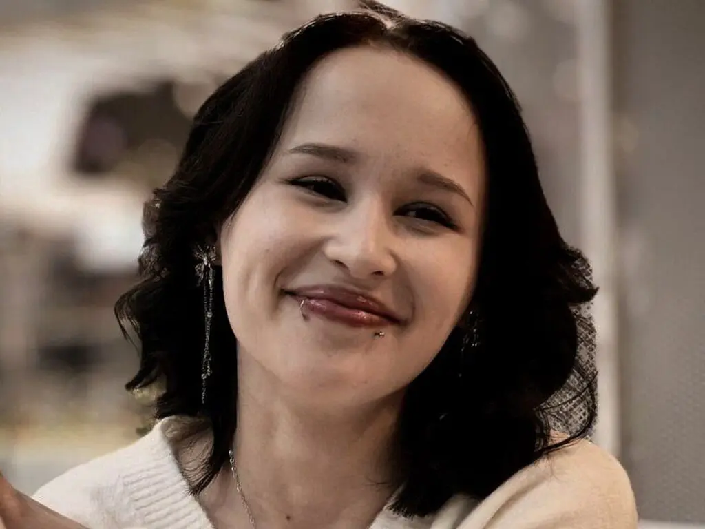 Smiling young woman with dark wavy hair and lip piercings wearing a cozy white sweater in a bright indoor setting.