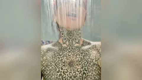 Woman with intricate black henna floral tattoos on her neck and shoulders in a soft, ethereal setting.