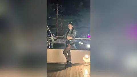 Stylish woman in a grey crop top and black shorts posing on a boat deck at night under a cloudy sky.