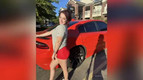 Smiling woman in a grey t-shirt and red shorts posing next to a bright red car in a sunny residential driveway.